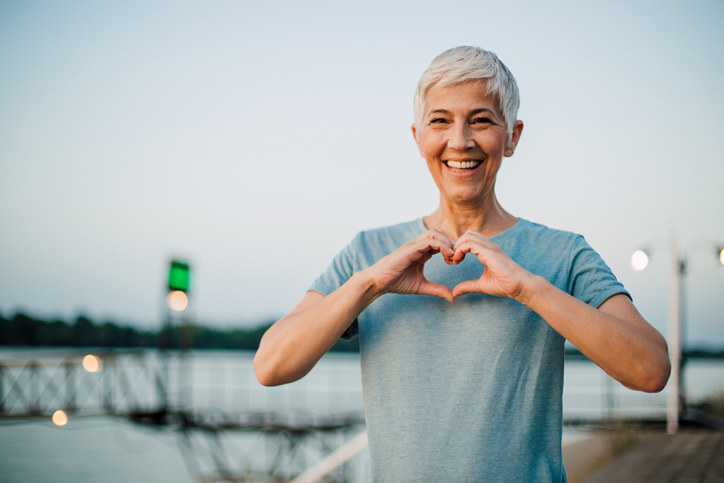 Portrait of a happy senior woman making a heart shape with her hands after exercising on the riverbank.