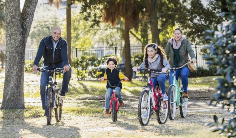 An Hispanic family with two children riding bicycles together in a city park on a sunny autumn day. The little 6 year old boy has a big grin on his face, riding in the middle between his parents and 10 year old sister.