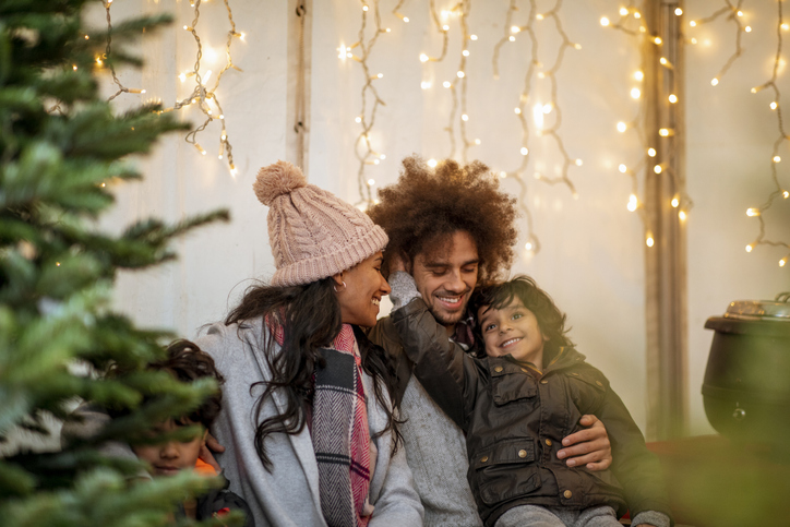 A multi-ethnic family at a Christmas tree farm together in Newcastle-Upon-Tyne. They are all sitting down in a gazebo while embracing, and looking at each other.