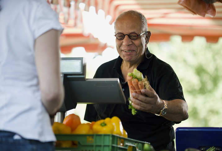 Mature man purchases a bundle of carrots at the checkout counter of an outdoor market. Horizontal shot.