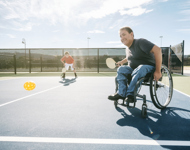 Two young men playing the game of pickleball on a court. One of the men is in a wheelchair. Horizontal composition with sunlight behind the players.