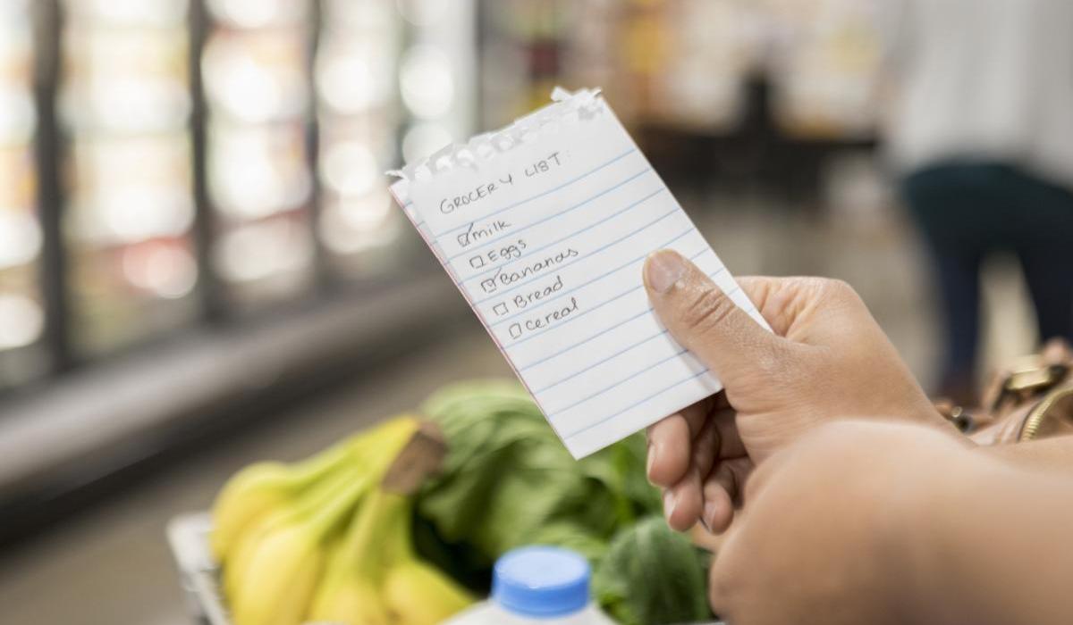In this point of view photograph, an unrecognizable person, with only hands showing, holds a paper shopping checklist over a full shopping cart. The customer is standing in the refrigerated foods aisle.