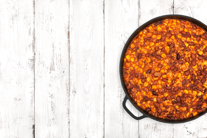 Hot chili con carne in a cast iron pan on light wooden background, top view