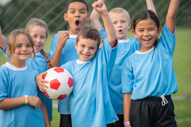 A multi ethnic group of elementary age children in a blue soccer team. They are dressed in a blue sports jersey and they have their hands in the air to celebrate their team's win.
