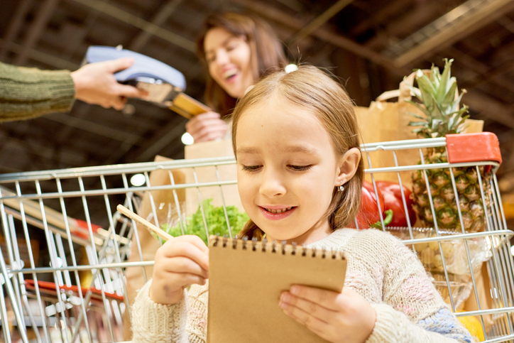 Warm-toned portrait of family doing grocery shopping in supermarket, focus on smiling little girl holding shopping list standing by cart