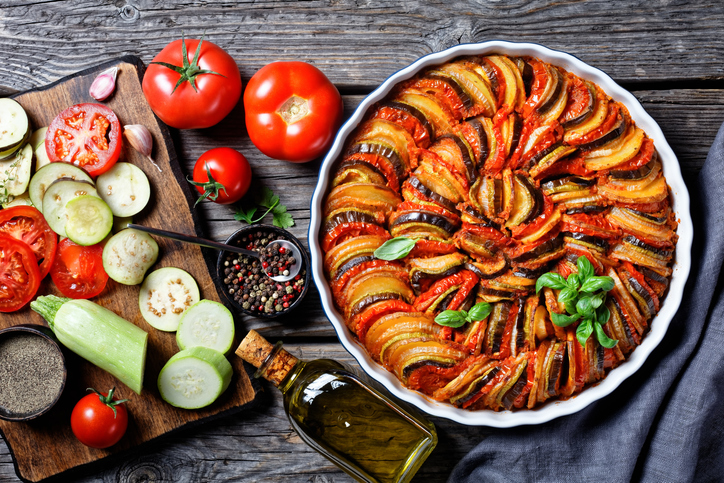 ratatouille, vegetable stew of sliced eggplant, zucchini, onion and potato with tomato sauce, ingredients at the background, french cuisine, horizontal view from above, flat lay