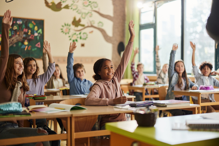 Children with raised hands sitting at desks in a classroom.