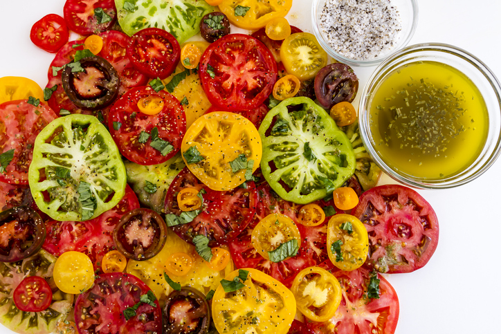 Slices of vine ripe heirloom tomato varieties with fresh basil, salt, pepper, olive oil and fresh herbs on white background