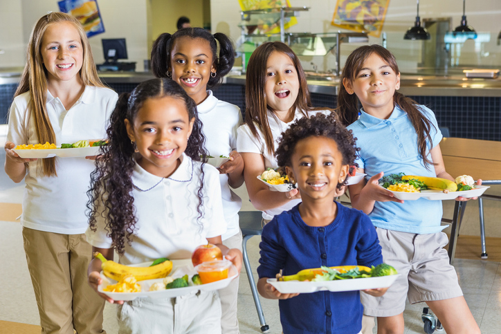 Group of children smiling and holding school lunch trays in a cafeteria.