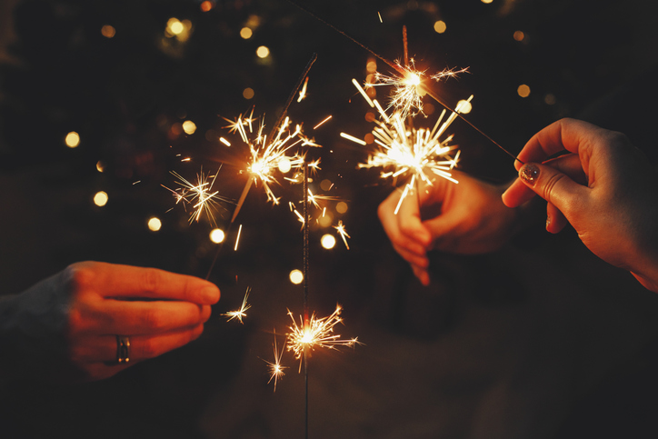 Happy New Year! Friends celebrating with burning sparklers in hands against christmas tree lights in dark room. Hands holding fireworks on background of stylish decorated illuminated tree.