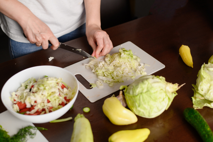 Woman prepares a vegan salad and cuts white cabbage. Close-up of a woman hands chopping white cabbage on a cutting board on the kitchen table.