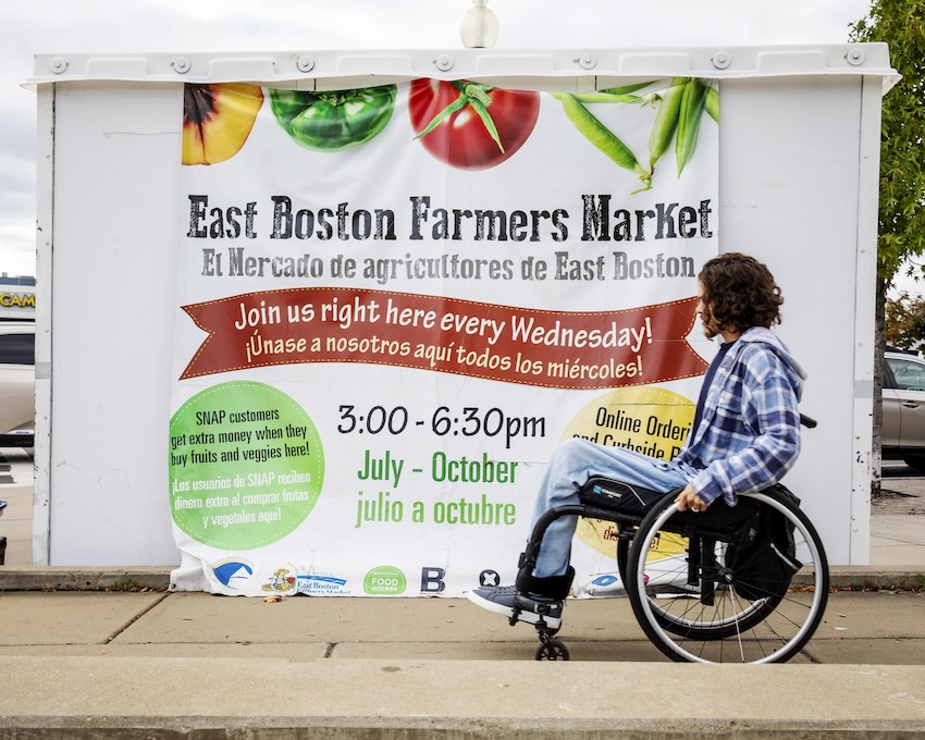 Young man in wheelchair rolls by a sign for a farmers' market