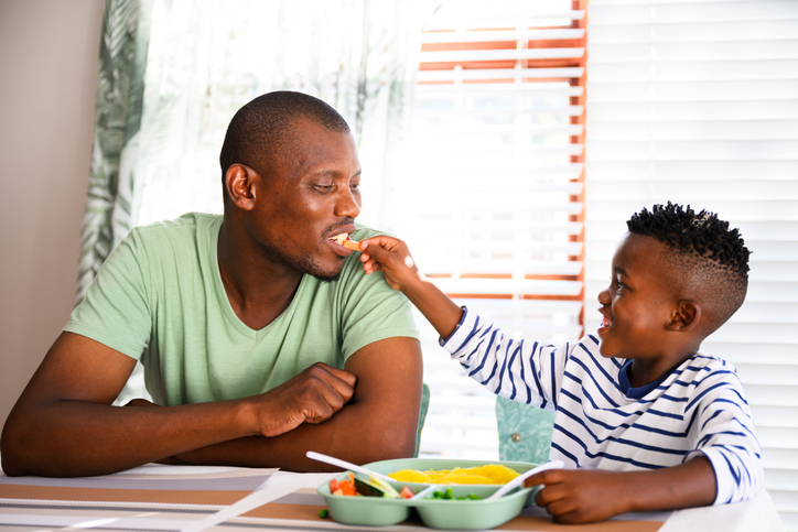 Father sitting with son eating vegetables for lunch. Little boy feeds his father a carrot sharing his healthy lunch