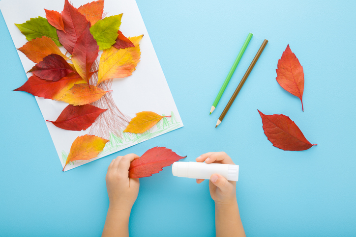 Little child hands gluing and creating tree shape from colorful leaves on white paper. Light blue table background. Pastel color. Making autumn decorations. Point of view shot. Closeup. Top down view.