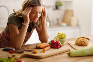 Mid adult female chef holding her head in pain while preparing a healthy salad in the kitchen.