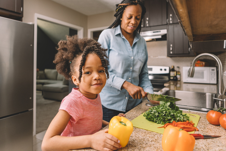 Little girl looking towards camera while her mom cuts vegetables. Healthy cooking with vegetables.