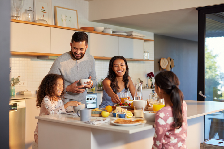 Hispanic parents and smiling little daughters enjoying a cheerful breakfast together in the kitchen. Mixed race mother and father sharing a joyful morning meal around the kitchen island with their cute little girls. Multiethnic family starting their day with healthy meal in their cozy home.