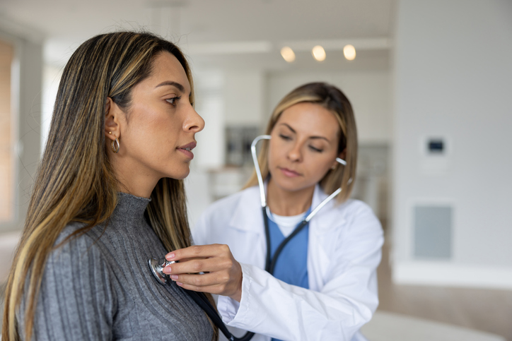 Latin American doctor on a house call performing a medical exam and listening to her heart with a stethoscope - healthcare and medicine concepts