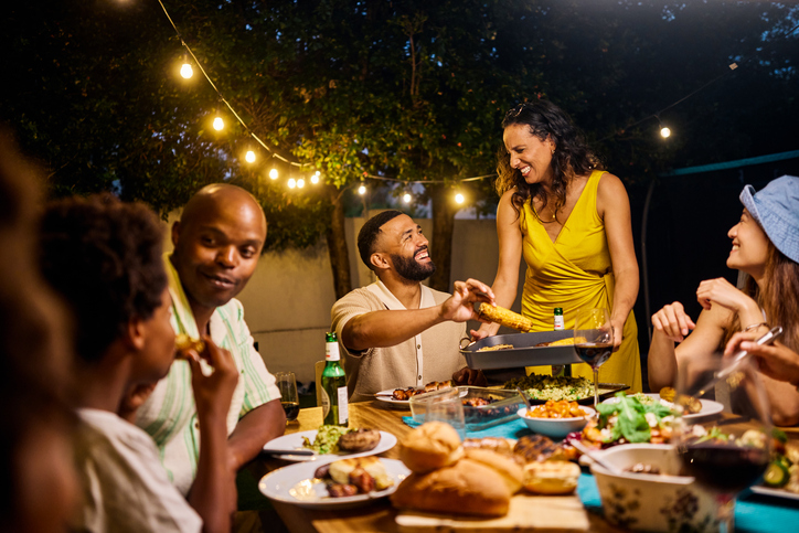 Family and friends dining at an outdoor table under string lights by the pool. Evening social gathering with food and drinks. Summer celebration concept. Medium shot.
