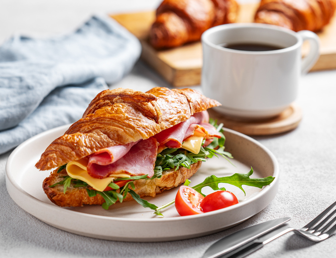 Croissant sandwich with ham, cheese, tomato and arugula on a plate on a white background with a cup of coffee, fresh pastry and morning light close up. Concept for a healthy breakfast or brunch.