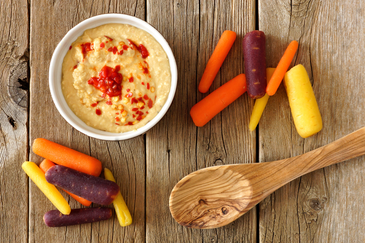 Baby rainbow carrots with hummus dip and spoon, overhead view on a rustic wooden background